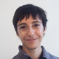 Portrait of Ennis Rook Bashe smiling, with short dark hair and subtle glitter makeup, wearing a grey collared shirt.