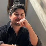 Tom Gilvear seated at a table indoors, wearing a black shirt and resting his hand against his chin.
